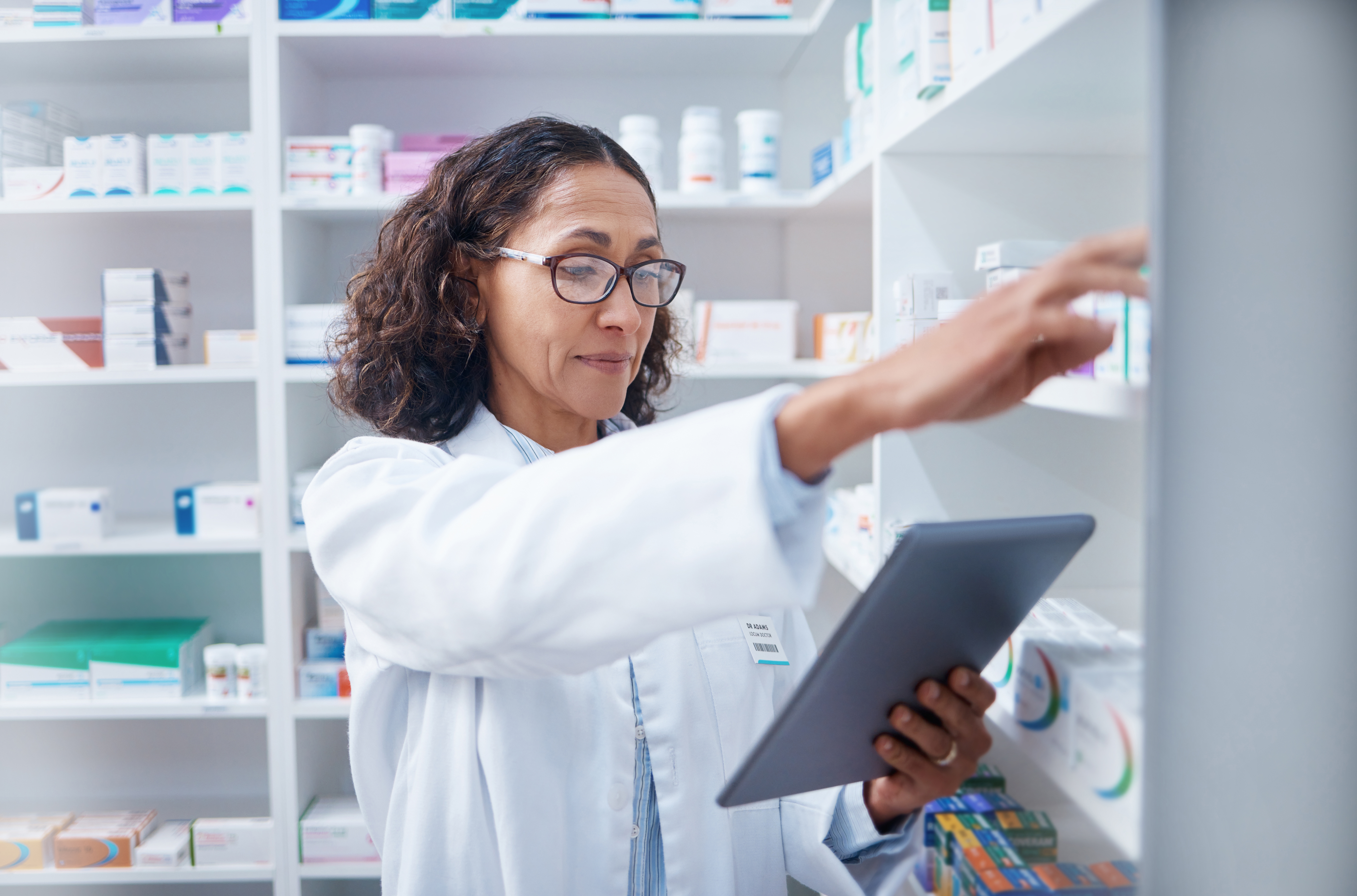 Independent pharmacist reviewing inventory on a tablet while organizing medications on pharmacy shelves.